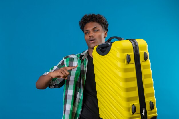 Young african american traveler man holding suitcase pointing with finger to it looking aside smiling confident standing over blue background