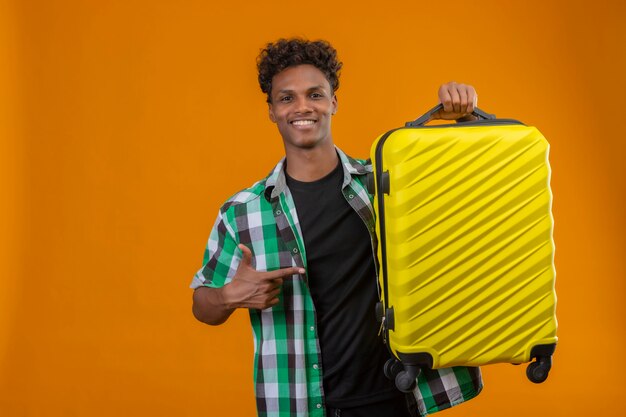 Young african american traveler man holding suitcase pointing with finger to it looking at camera smiling cheerfully standing over orange background