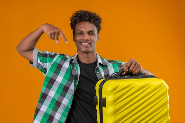 Young african american traveler man holding suitcase pointing with finger to it smiling cheerfully