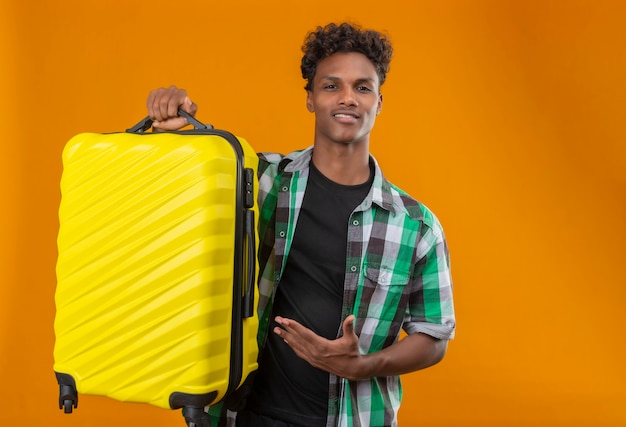 オレンジ色の背景に自信を持って肯定的で幸せな立っている笑顔のカメラを見て彼の手の腕を提示するスーツケースを持って若いアフリカ系アメリカ人旅行者男