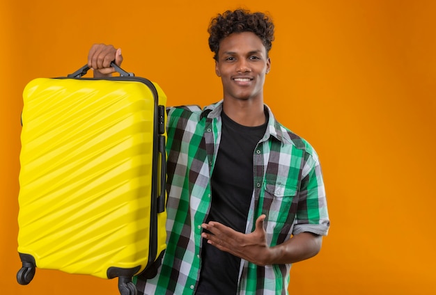 Young african american traveler man holding suitcase presenting with arm of his hand looking at camera smiling confident, positive and happy