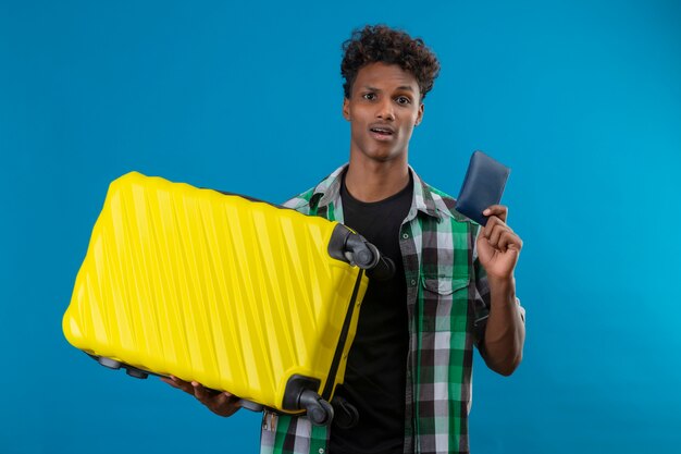 Молодой афро-американский путешественник мужчина держит чемодан и бумажник, глядя в камеру, смущенный и удивленный