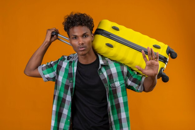 Young african american traveler man holding suitcase with open hand, making stop sign, defense gesture over orange backghround