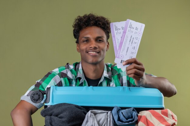 Young african american traveler man standing with suitcase full of clothes holding air tickets looking at camera smiling cheerfully positive and happy over green background