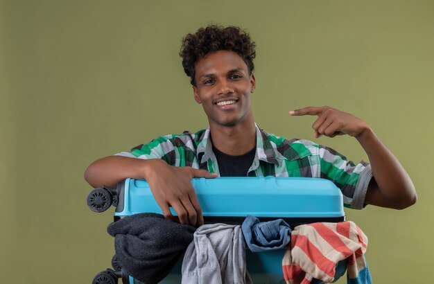 Young african american traveler man standing with suitcase full of clothes looking at camera happy and positive smiling pointing with finger to the side over green background