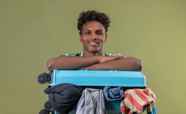Молодой афро-американский путешественник, стоящий с чемоданом, полным одежды, глядя в камеру, улыбается позитивно и счастливо на зеленом фоне