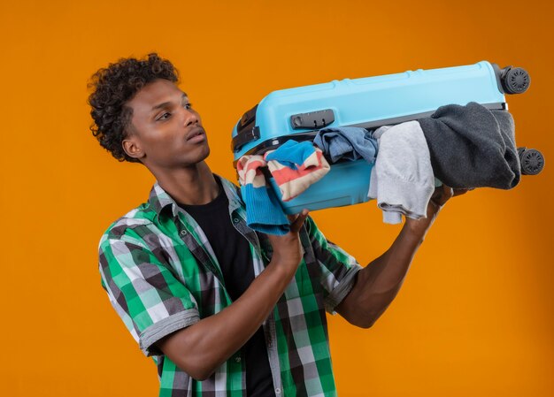 Молодой афро-американский путешественник стоит с чемоданом, полным одежды, выглядит смущенным на оранжевом фоне