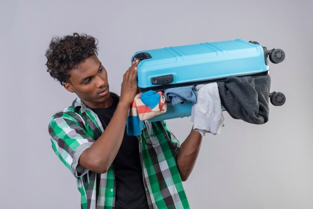 Молодой афро-американский путешественник, стоящий с чемоданом, полным одежды, выглядит смущенным на белом фоне
