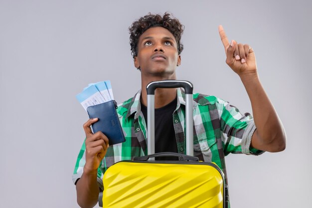 スーツケースを持って立っている若いアフリカ系アメリカ人旅行者男航空券を探していると白い背景の上の深刻な顔で指を上向き