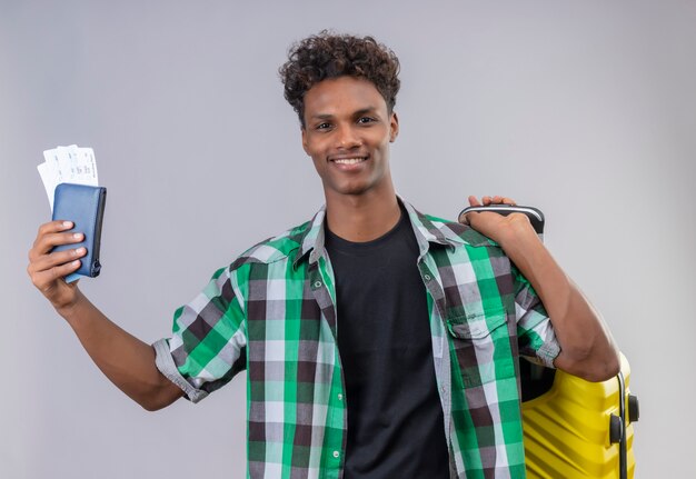 Young african american traveler man standing with suitcase holding air tickets smiling cheerfully positive and happy over white background