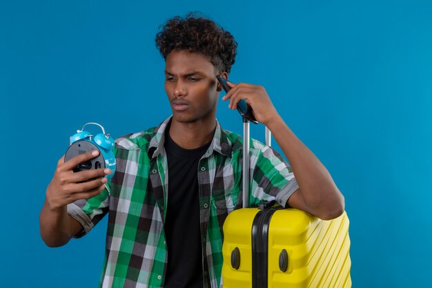 青い背景に心配してそれを見て目覚まし時計を保持しているスーツケースを持って立っている若いアフリカ系アメリカ人旅行者男