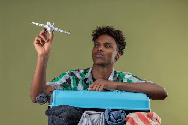 緑の背景の上の顔に真剣な自信のある表情でそれを見ておもちゃの飛行機を保持しているスーツケースを持って立っている若いアフリカ系アメリカ人旅行者男