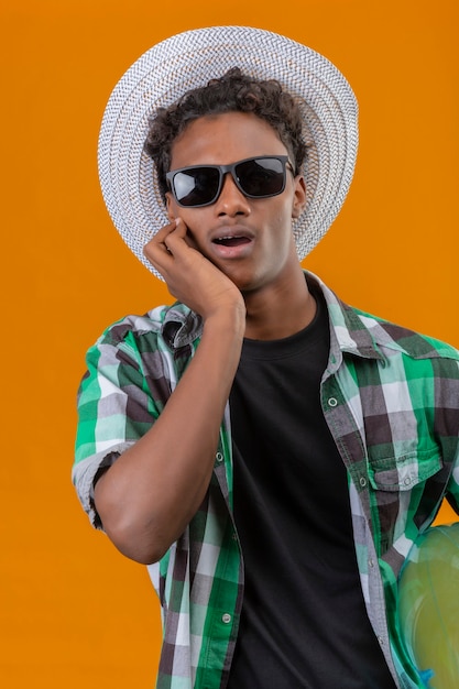 Young african american traveler man in summer hat wearing black sunglasses holding inflatable ring amazed and surprised looking at camera standing over orange background