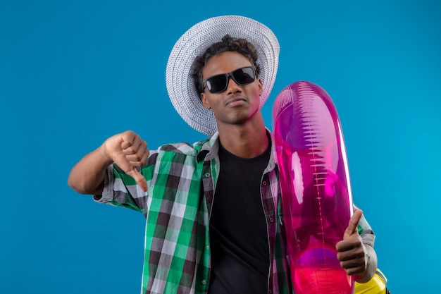 Young african american traveler man in summer hat wearing black sunglasses holding inflatable ring  displeased showing thumbs down standing over blue background