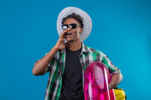 Young african american traveler man in summer hat wearing black sunglasses holding inflatable ring looking aside shouting or calling someone standing over blue background