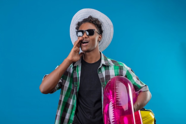 Young african american traveler man in summer hat wearing black sunglasses holding inflatable ring looking aside shouting or calling someone  