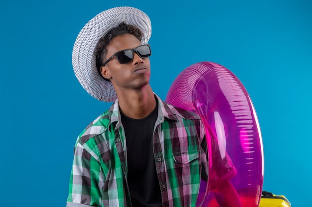 Молодой афро-американский путешественник в летней шляпе, одетый в черные солнцезащитные очки, держит надувное кольцо, глядя в сторону с уверенным серьезным выражением лица, стоящим на синем фоне