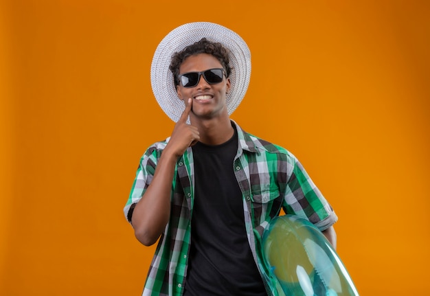 Young african american traveler man in summer hat wearing black sunglasses holding inflatable ring looking at camera with big smile on face pointing with finger to his smile standing over ora