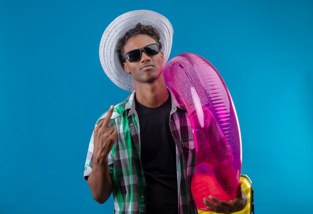 Young african american traveler man in summer hat wearing black sunglasses holding inflatable ring looking confident, satisfied , showing middle finger  