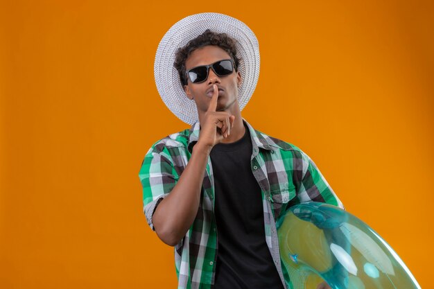 Young african american traveler man in summer hat wearing black sunglasses holding inflatable ring making silence gesture with finger on lips standing over orange background