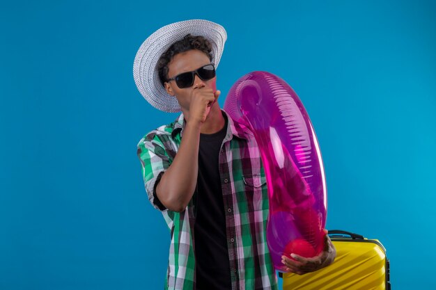 Young african american traveler man in summer hat wearing black sunglasses standing with suitcase holding inflatable ring coughing over blue background