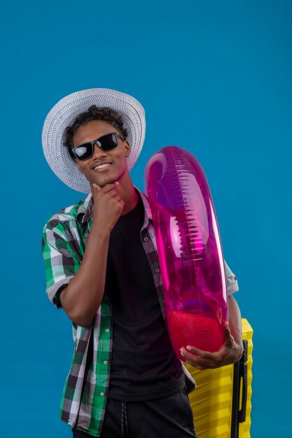 Young african american traveler man in summer hat wearing black sunglasses standing with suitcase holding inflatable ring looking at camera smiling cheerfully over blue background