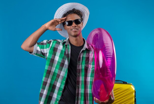 Young african american traveler man in summer hat wearing black sunglasses standing with suitcase holding inflatable ring saluting and smiling over blue background