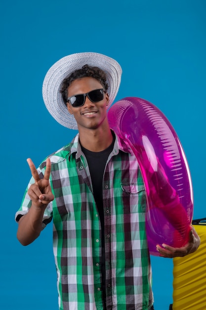 Молодой афро-американский путешественник в летней шляпе, одетый в черные солнцезащитные очки с чемоданом, держащим надувное кольцо, показывающий номер два или знак победы, улыбаясь веселый