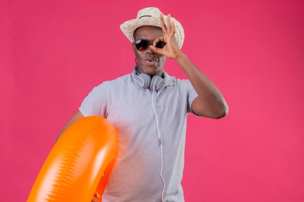 Young african american traveler man in summer hat with headphones around his neck wearing black sunglasses holding inflatable ring doing ok sing looking through this sing over pink b