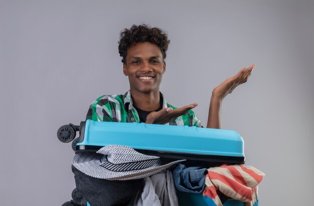 Young african american traveler man with suitcase full of clothes looking at camera smiling cheerfully presenting with arms of his hands standing over white background