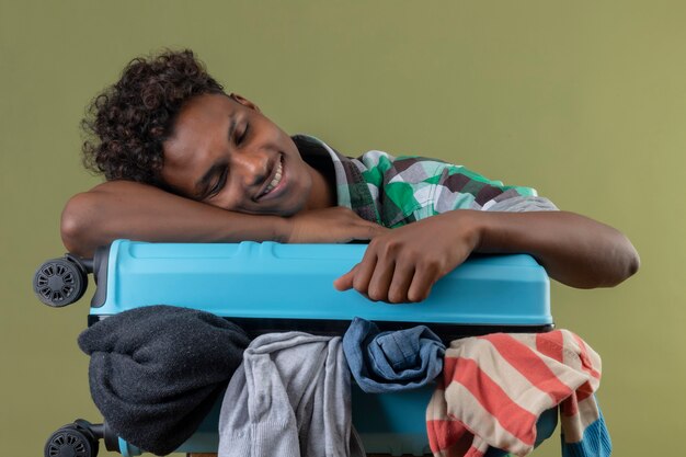 Young african american traveler man with suitcase full of clothes looking tired sleeping on it over green background