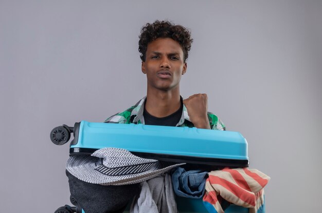 彼の成功を喜んで自信を持って見える拳を上げる服でいっぱいのスーツケースを持つ若いアフリカ系アメリカ人の旅行者の男