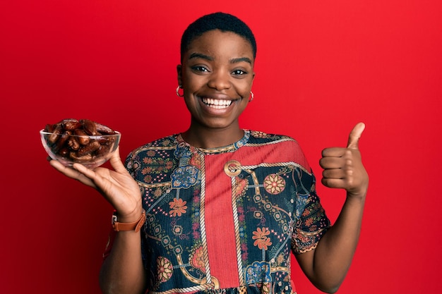Young african american woman holding bowl with dates smiling happy and positive, thumb up doing excellent and approval sign