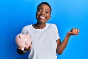Free photo young african american woman holding piggy bank screaming proud celebrating victory and success very excited with raised arm