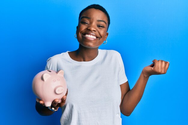 Young african american woman holding piggy bank screaming proud celebrating victory and success very excited with raised arm