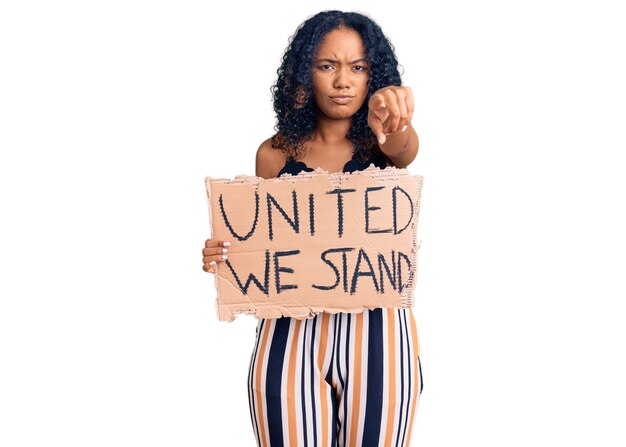Молодая африканская американка, держащаяся за единство, мы стоим баннер, указывая пальцем на камеру и на вас, уверенный жест выглядит серьезным