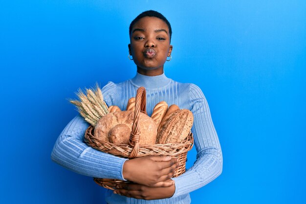 Free photo young african american woman holding wicker basket with bread puffing cheeks with funny face mouth inflated with air catching air