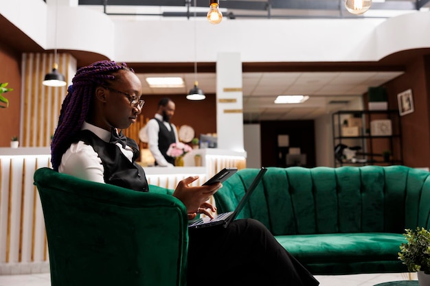 Молодая афроамериканская женщина-менеджер отеля сидит в приемной, используя смартфон и ноутбук, управляет бюджетом и заказывает принадлежности. Технологии в индустрии гостеприимства