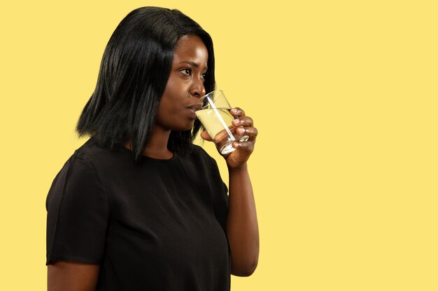 Young african-american woman isolated on yellow studio background, facial expression. Beautiful female half-length portrait. Concept of human emotions, facial expression. Drinking water and smiling.
