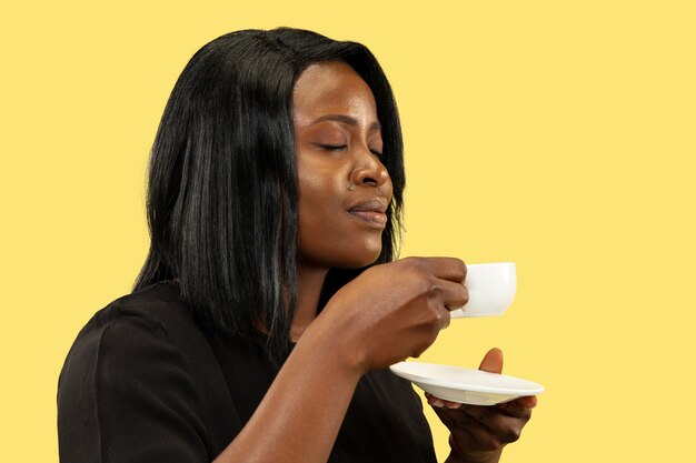 노란색 스튜디오 배경, 표정에 고립 된 젊은 아프리카 계 미국인 여자. 아름 다운 여성 절반 길이 초상화. 인간의 감정, 표정의 개념. 커피 마시기.