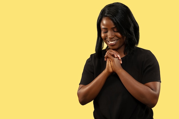 Молодая афро-американская женщина, изолированные на желтом фоне студии, выражение лица. Красивый женский поясной портрет. Понятие о человеческих эмоциях, выражении лица. Молиться и улыбаться.