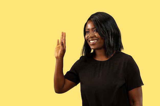 Young african-american woman isolated on yellow studio background, facial expression. Beautiful female half-length portrait. Concept of human emotions, facial expression. Showing the sign of bye.