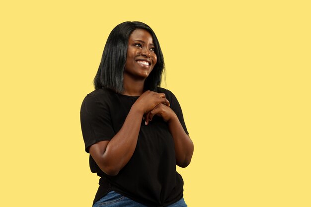 Young african-american woman isolated on yellow studio background, facial expression. Beautiful female half-length portrait. Concept of human emotions, facial expression. Standing and smiling.