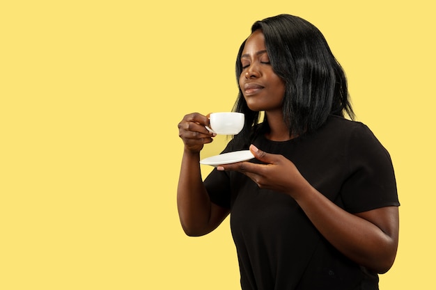 Young african-american woman isolated on yellow wall, facial expression. Beautiful female half-length portrait. Concept of human emotions, facial expression. Enjoying of drinking coffee.