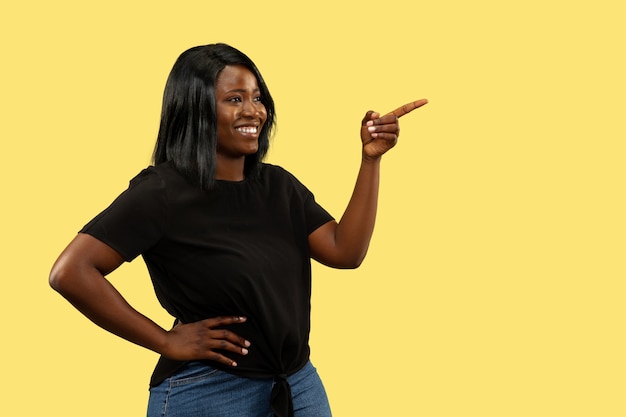 Young african-american woman isolated on yellow wall, facial expression. Beautiful female half-length portrait. Concept of human emotions, facial expression. Pointing and smiling.