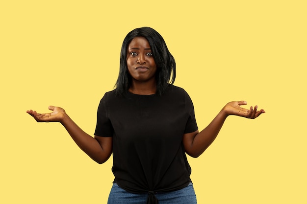 Young african-american woman isolated on yellow wall, facial expression. Beautiful female half-length portrait. Concept of human emotions, facial expression. Uncertainty, confusion.