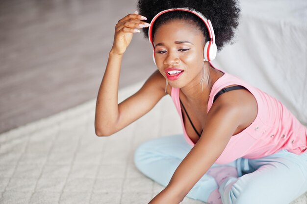 ピンクの一重項の若いアフリカ系アメリカ人女性は彼女の白い部屋でイヤホンで音楽を聴きます