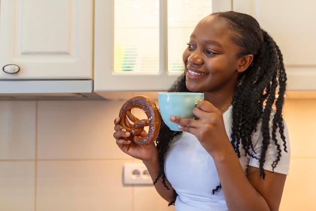 Young African American woman sitting on a kitchen desk and enjoying her pretzel with cup of worm milk