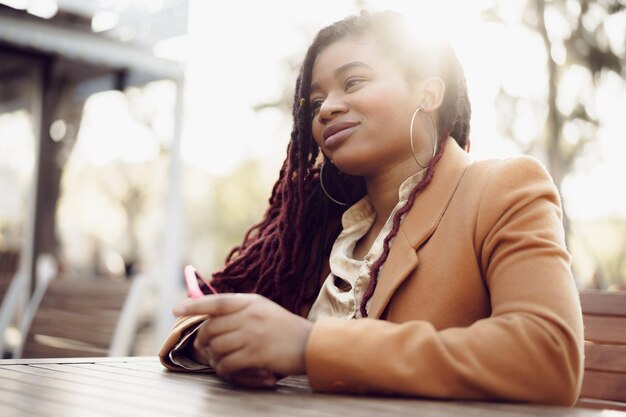 Молодая афро-американская женщина сидит в уличном кафе и держит смартфон в руках