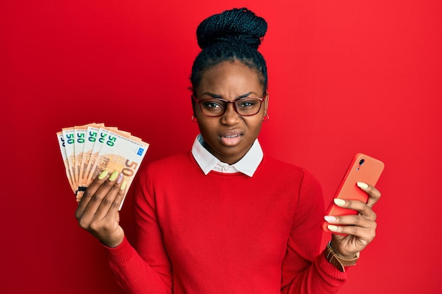 ユーロ紙幣を持つスマートフォンを使用する若いアフリカ系アメリカ人の女性は、無知で混乱した表情をしています。疑いの概念。